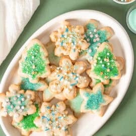 Vegan gluten free sugar cookies decorated with green and blue sprinkles piled on a white platter on a green surface.