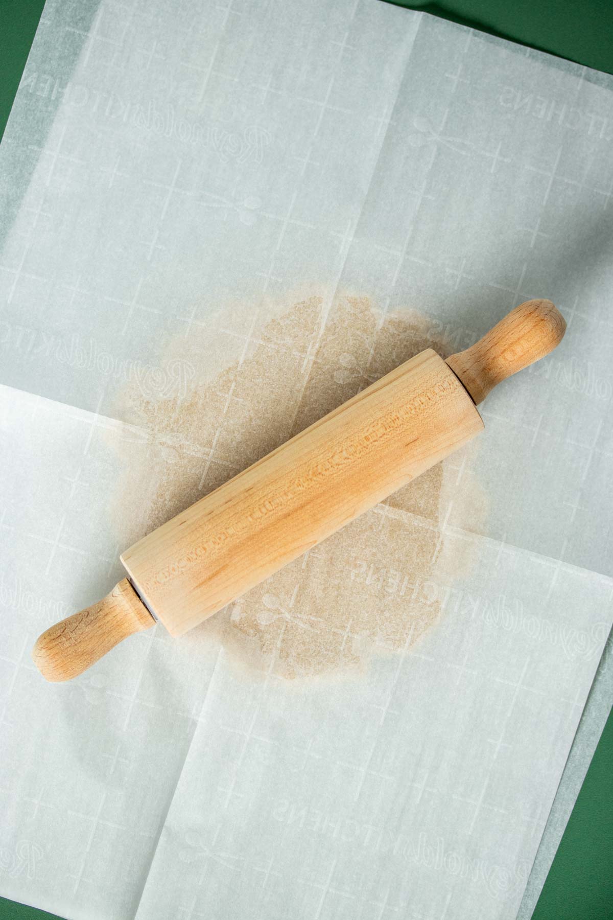 A rolling pin on top of a piece of parchment paper with cookie dough under it.