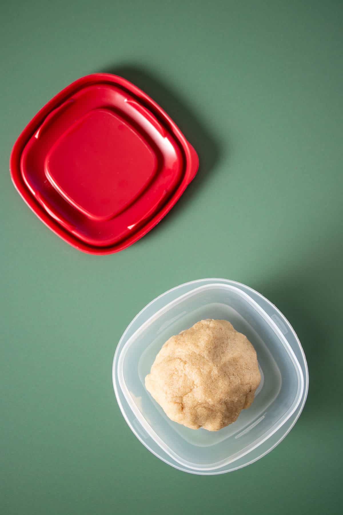 A ball of sugar cookie dough in a plastic square container on a green surface with a red lid next to it.