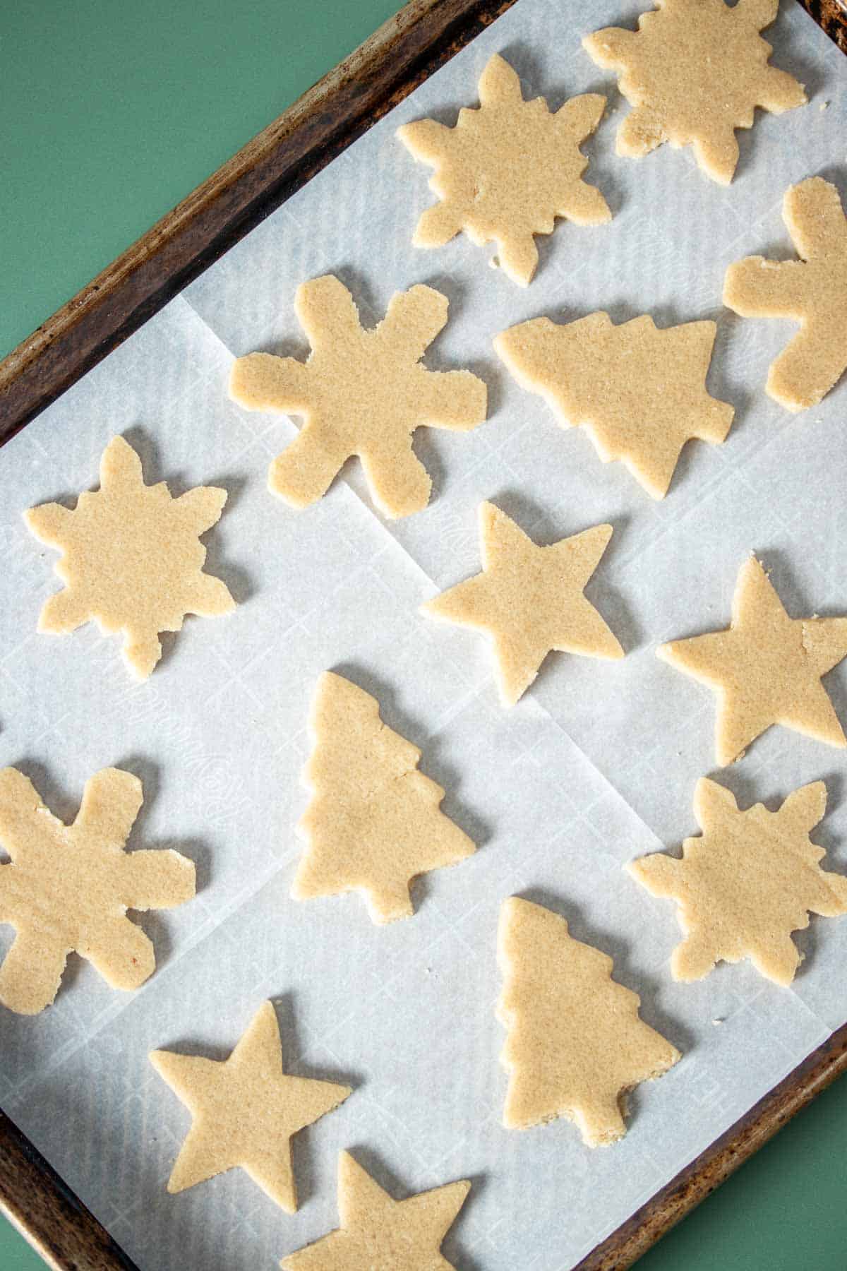 Unbaked sugar cookies in holiday shapes sitting on a parchment lined baking sheet.