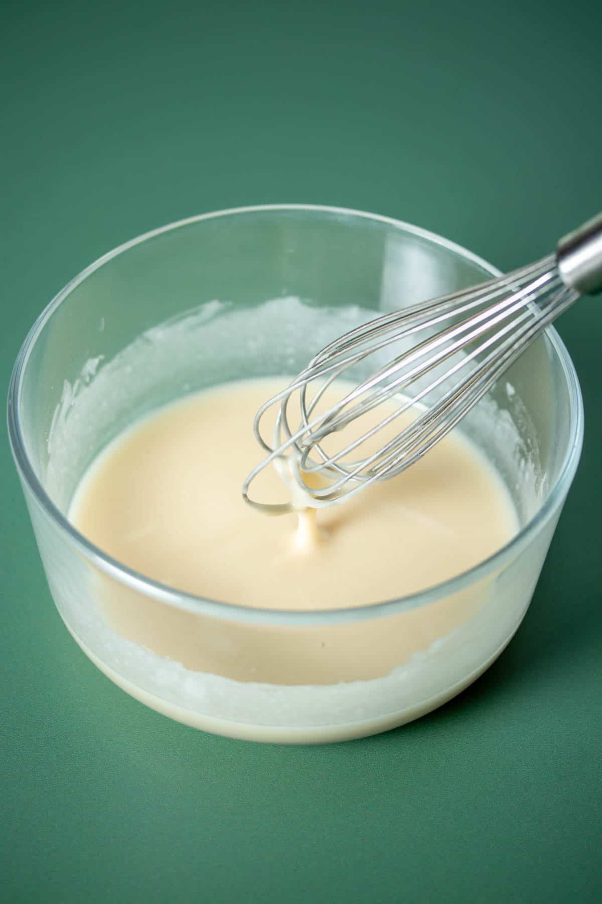 A whisk mixing white icing in a glass bowl on a green surface.