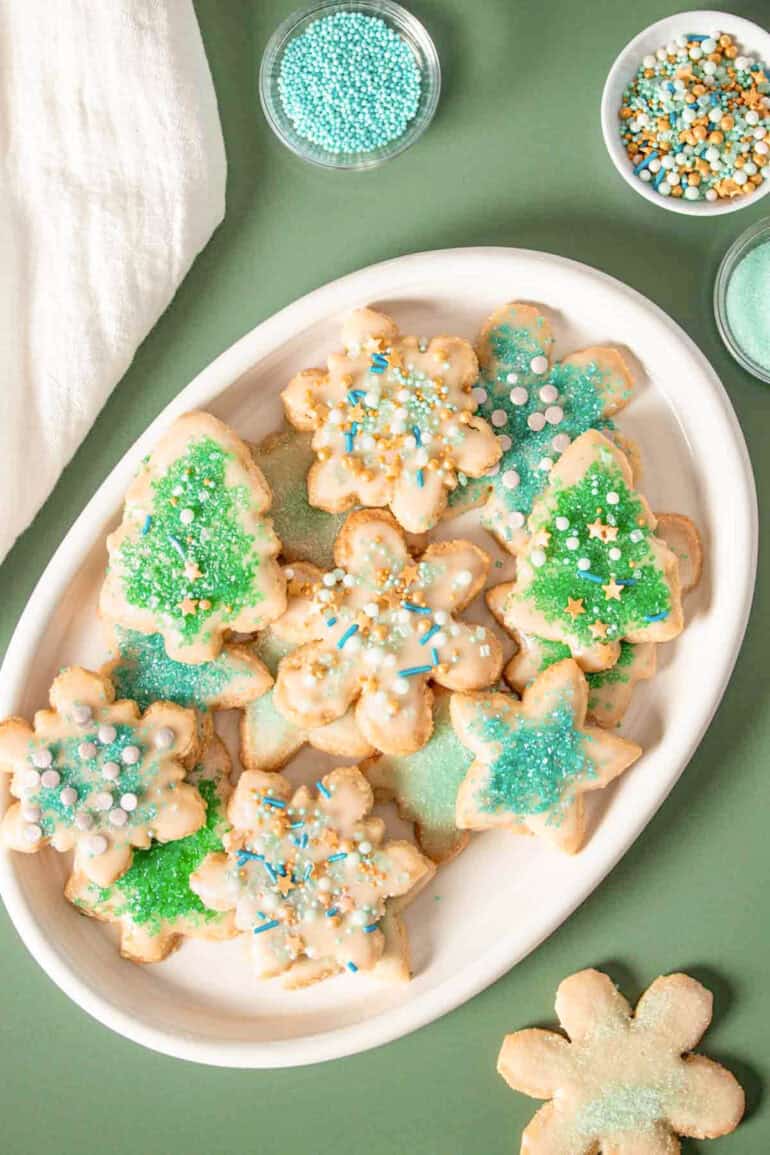 Vegan gluten free sugar cookies decorated with green and blue sprinkles piled on a white platter on a green surface.