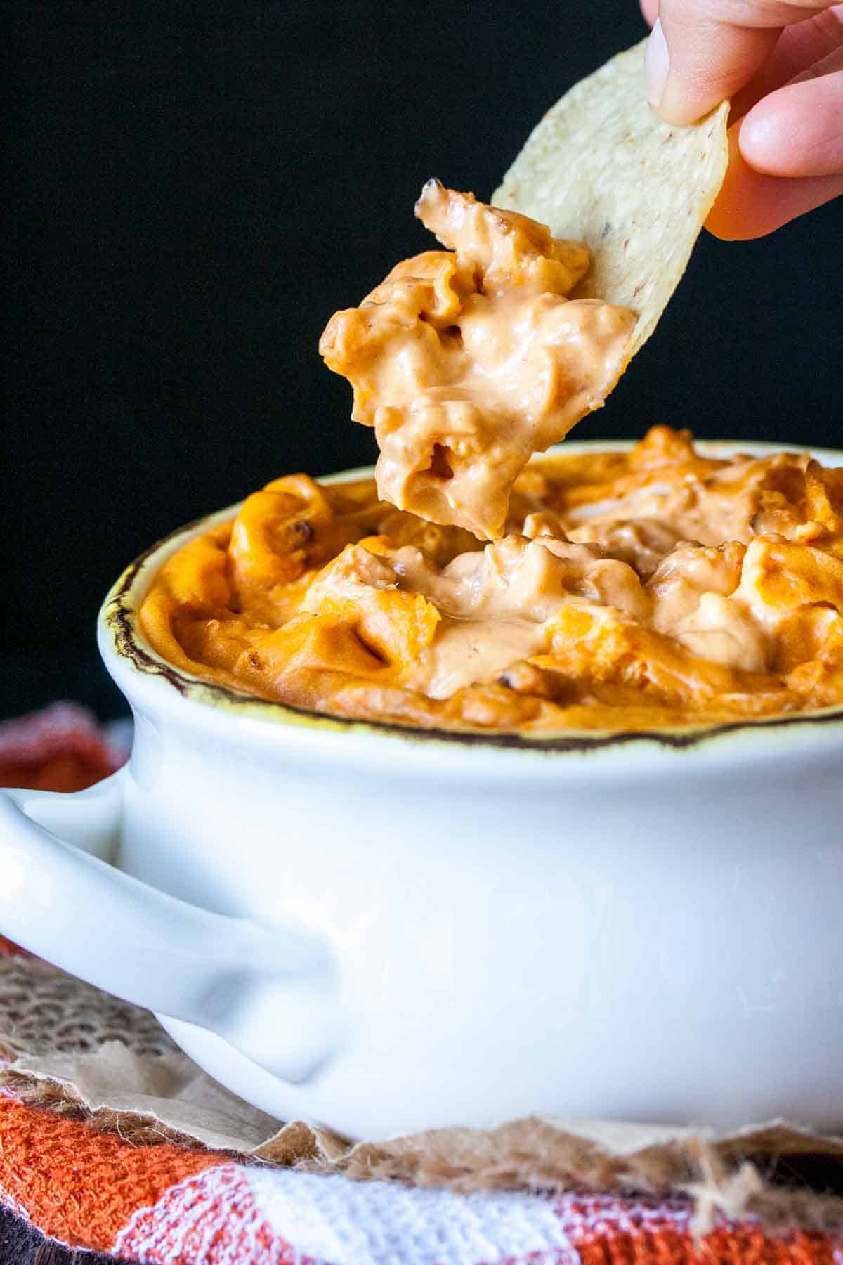 Chip taking a bite from a bowl of chunky vegan buffalo dip with cauliflower pieces.