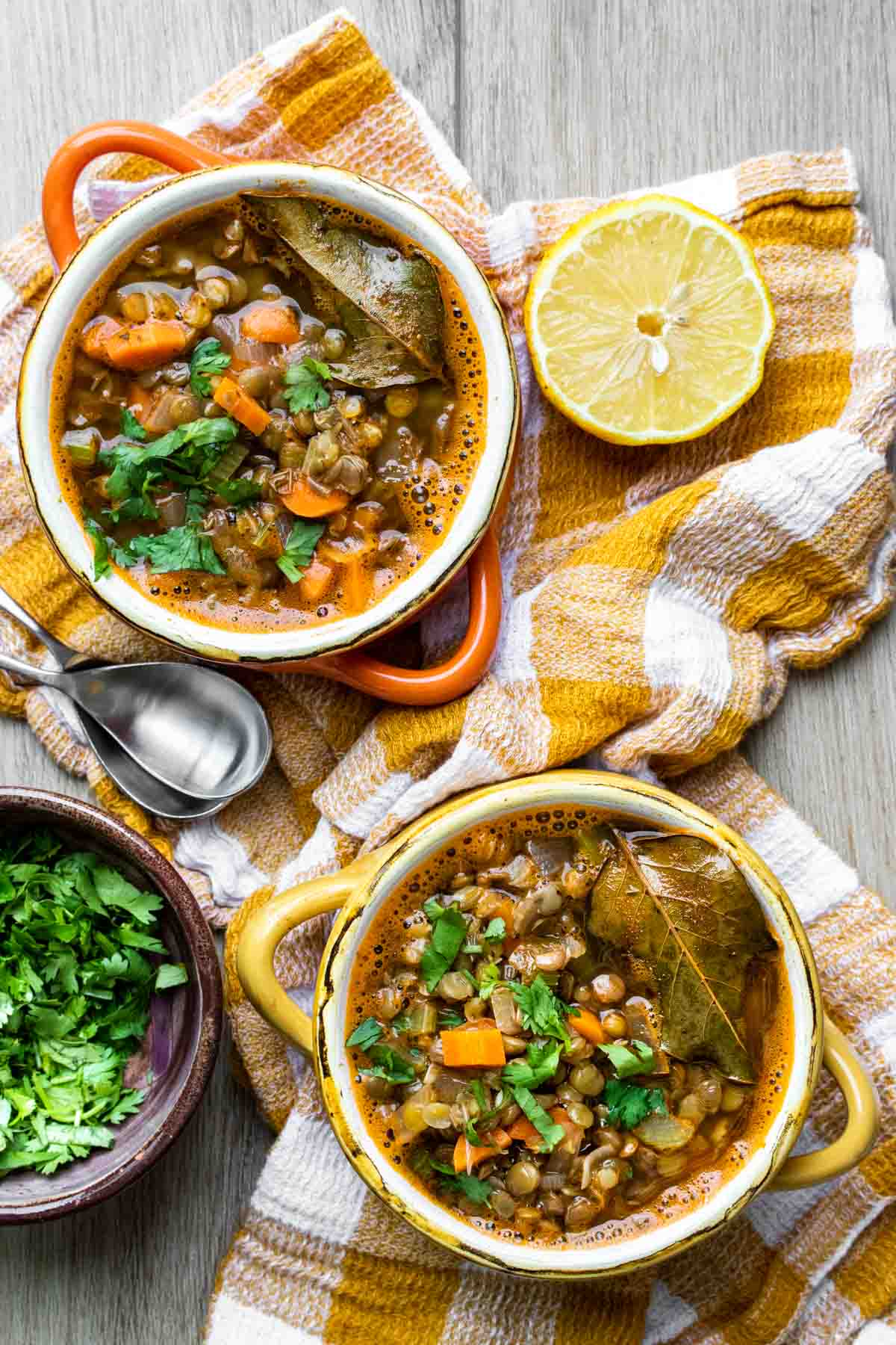 An orange and a yellow soup bowl filled with Greek lentil soup on a yellow checkered towel next to toppings.