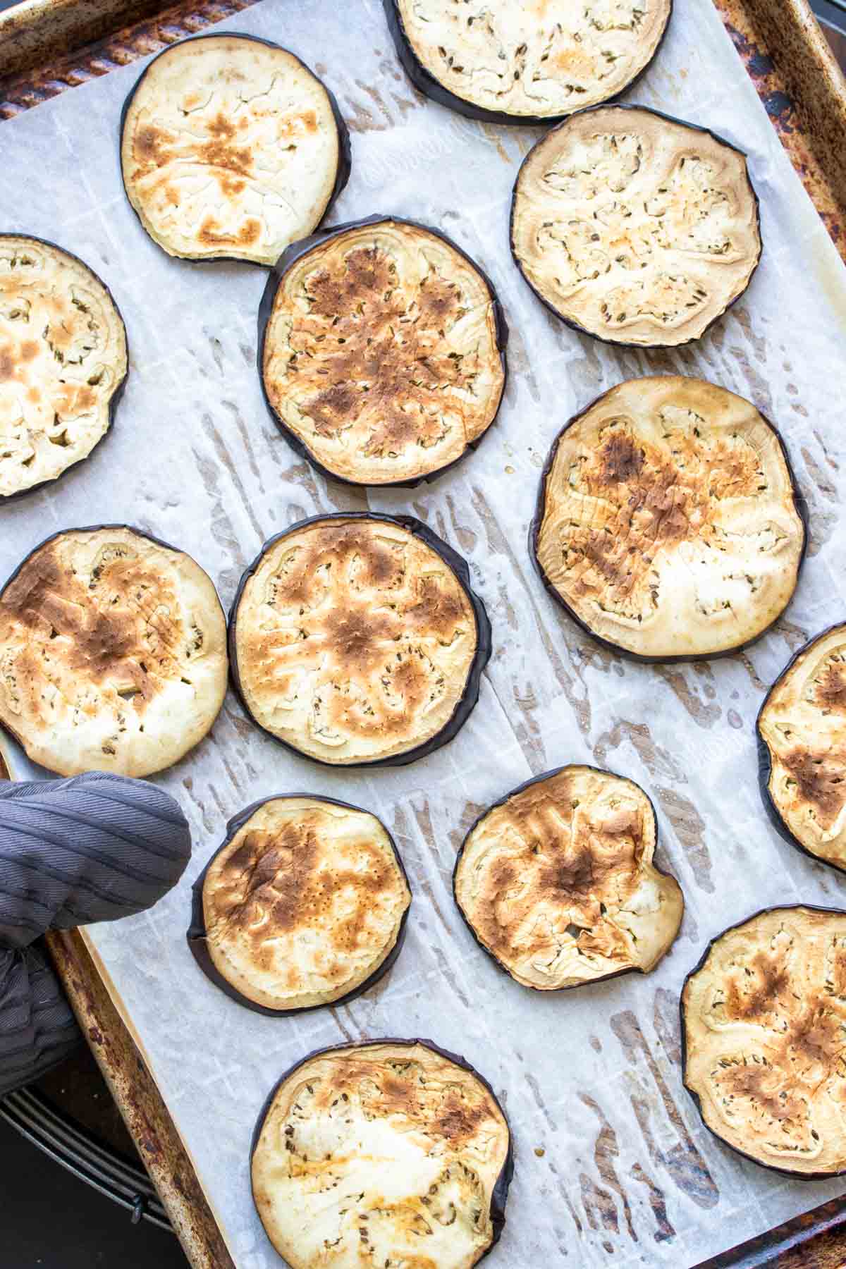 A grey oven mitt holding a baking pan with parchment and round slices of baked eggplant.