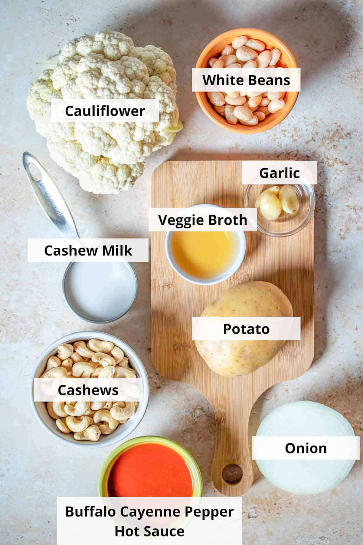 Top view of a tan surface with ingredients for a vegan buffalo chicken dip recipe: potatoes, cauliflower, onion, hot sauce, broth, milk, beans, nuts and garlic on it.