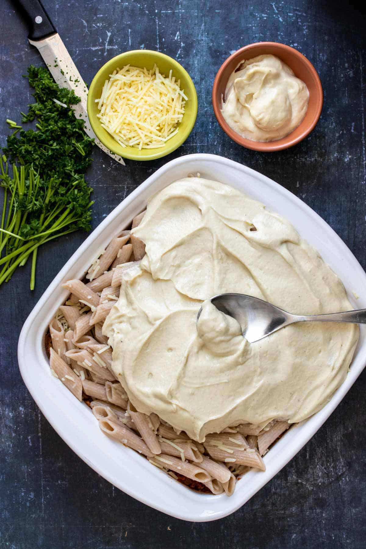 A spoon spreading a Bechamel sauce over penne pasta in a white baking dish next to chopped parsley and cheese and more bechamel in bowls