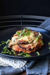 Black plate with a piece of vegan gluten-free eggplant parmesan topped with sliced basil.