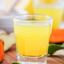 Homemade lemon ginger turmeric wellness shots served in a shot glass on a wooden cutting board.