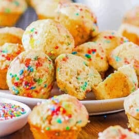 Birthday cake muffins, stacked on a white plate with sprinkles on the side.