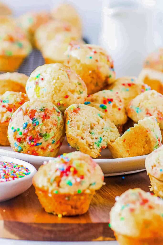 Birthday cake muffins, stacked on a white plate with sprinkles on the side.