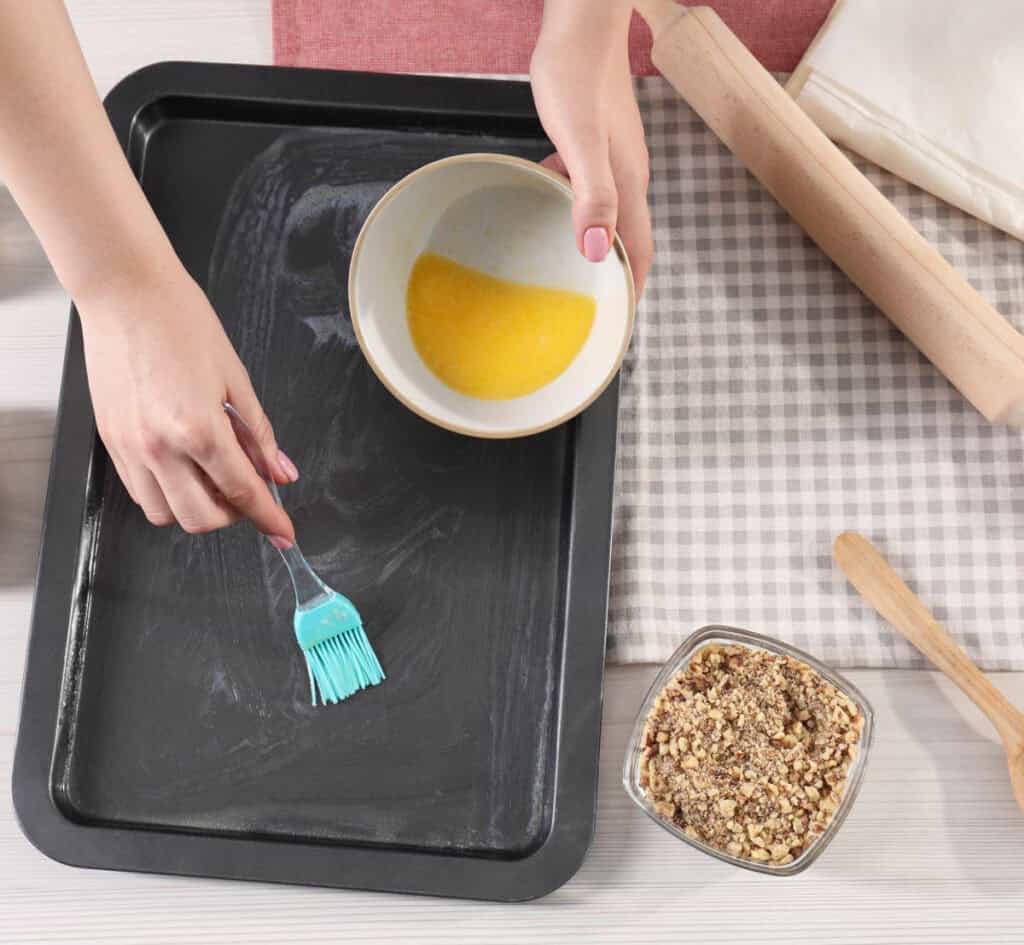 Making healthy baklava. Woman buttering baking pan at white wooden table, closeup.