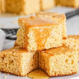 Healthy cornbread squares drizzled with honey, stacked on a white plate.