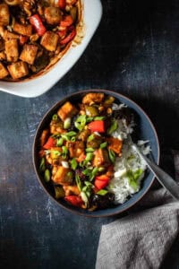 Vegan kung pao tofu in a dark blue bowl with white rice and sprinkled with green onions next to a grey towel, with a fork in the bowl.