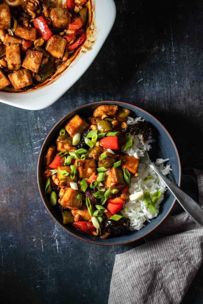 Vegan kung pao tofu in a dark blue bowl with white rice and sprinkled with green onions next to a grey towel, with a fork in the bowl.