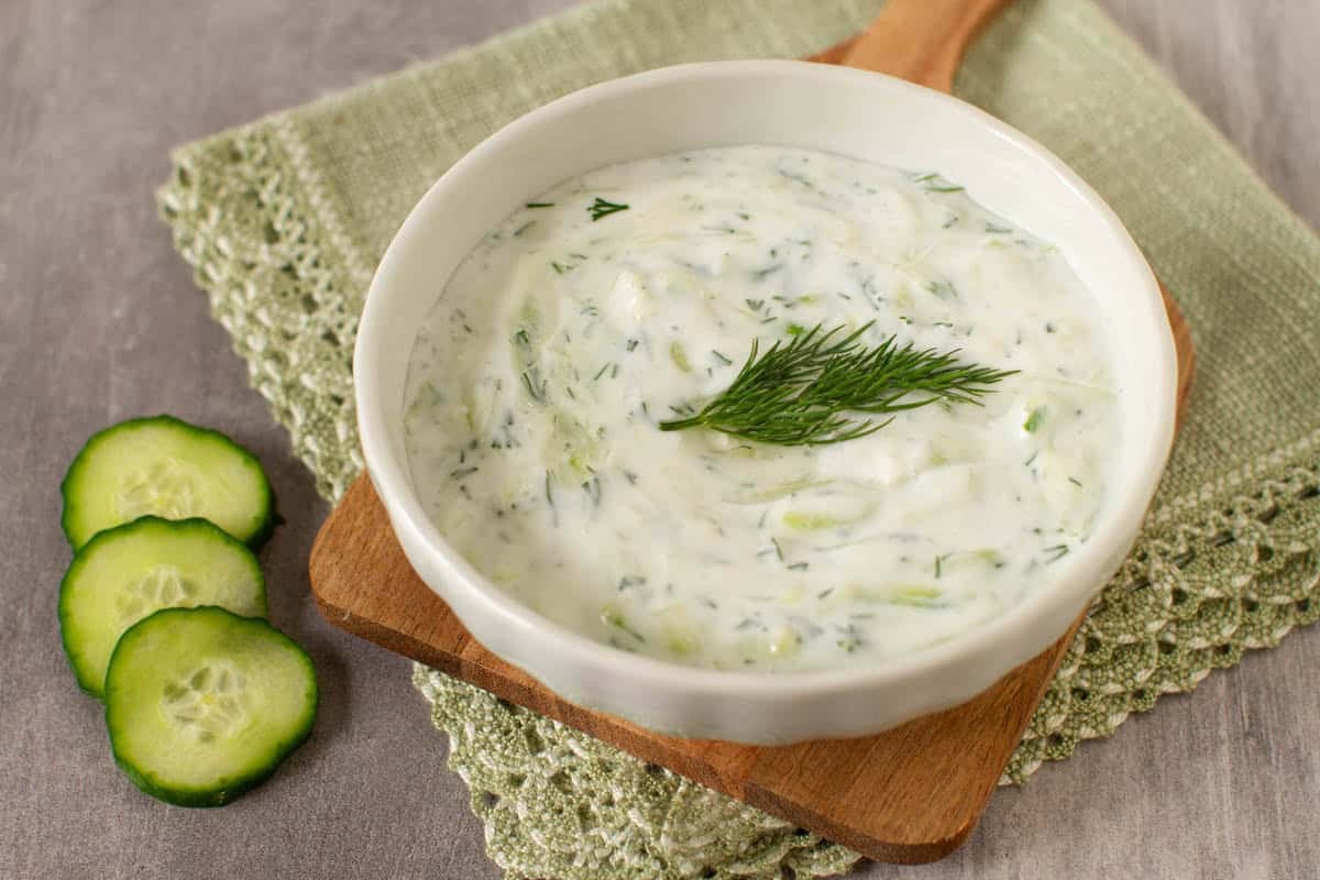 Vegan tzatziki sauce in a white bowl topped with fresh dill and sliced cucumbers on the side.