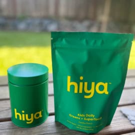 Container and pouch of Hiya Vanilla Greens + Superfoods for Kids on a wood deck against a nature background.