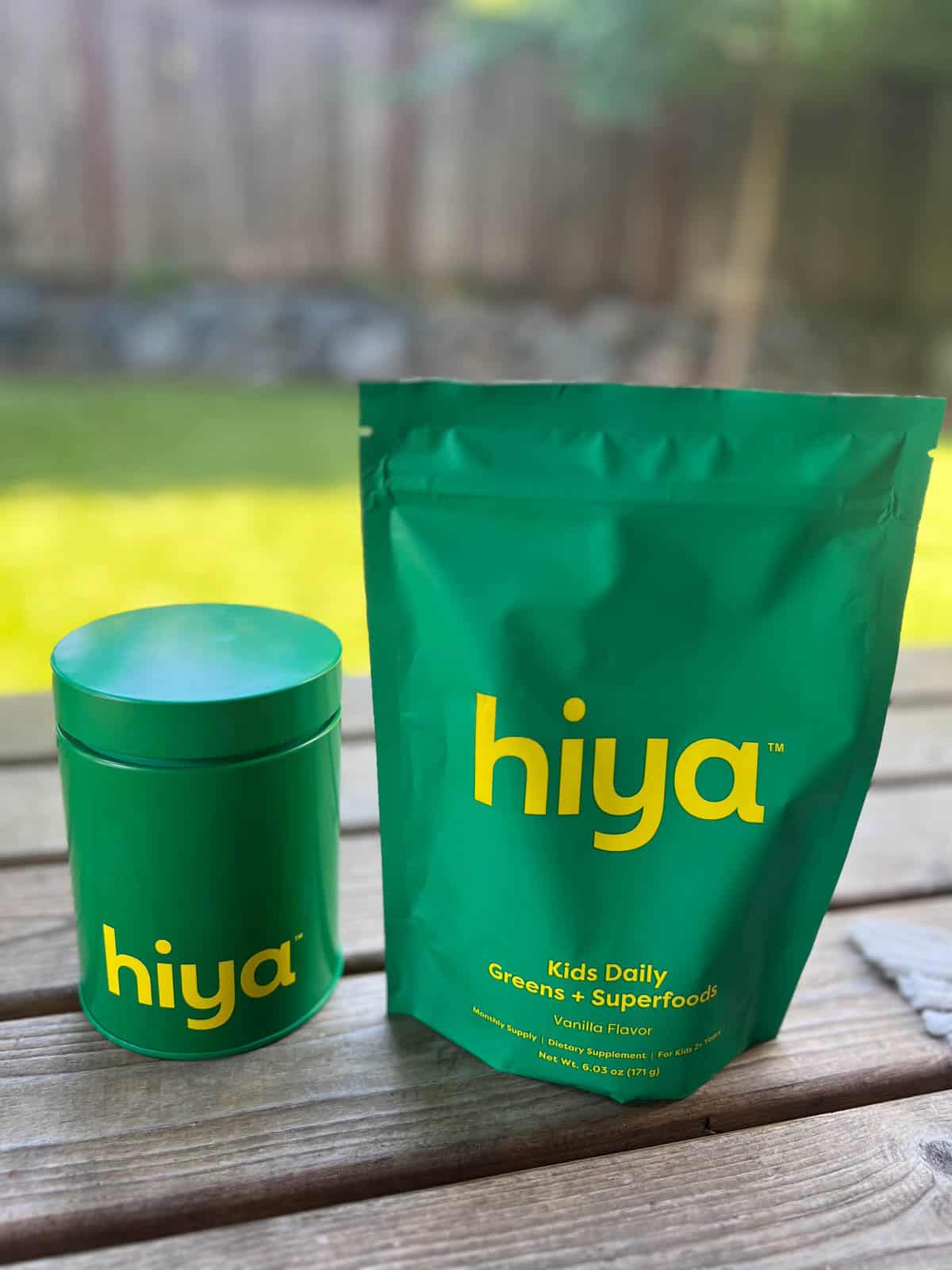 Container and pouch of Hiya Vanilla Greens + Superfoods for Kids on a wood deck against a nature background.