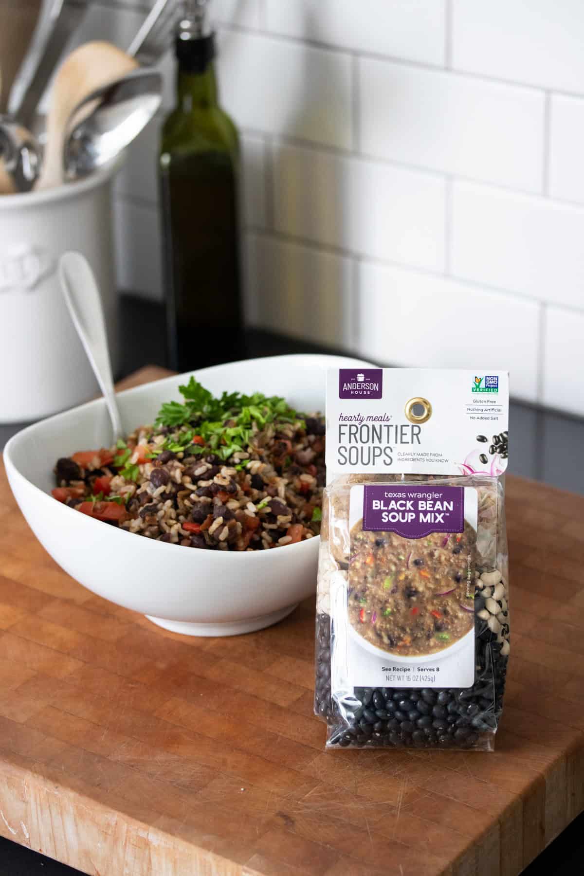 A bag of black bean soup mix sitting on a wooden surface in front of a white bowl with a black bean and rice salad in it.