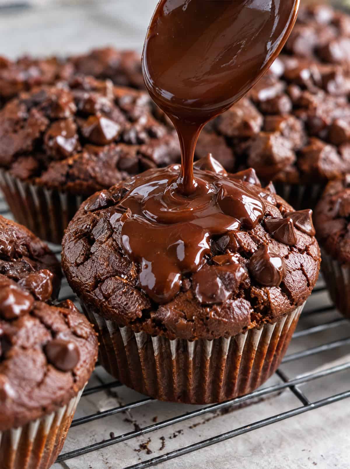 Chocolate sauce being drizzled over chocolate sweet potato muffins on a wire rack.